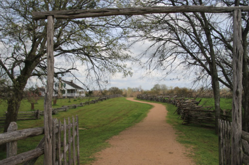 A rustic wooden gate opens to a dirt path lined with trees, leading to a house in the distance.