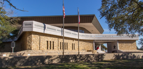 Exterior view of a modern stone building with flags, featuring a star emblem and surrounded by trees.