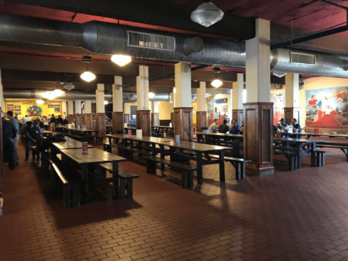 A spacious dining area with wooden tables and benches, featuring a few patrons and colorful wall murals.