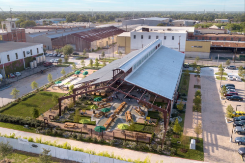 Aerial view of a large, modern building with a metal roof, surrounded by greenery and outdoor seating areas.
