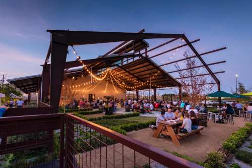 A lively outdoor venue with string lights, people socializing, and greenery, set against a twilight sky.