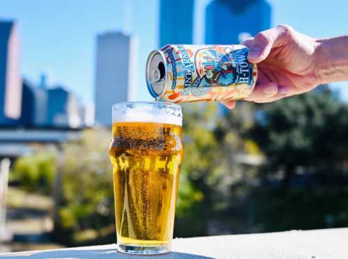 A hand pours a can of beer into a glass, with a city skyline in the background.