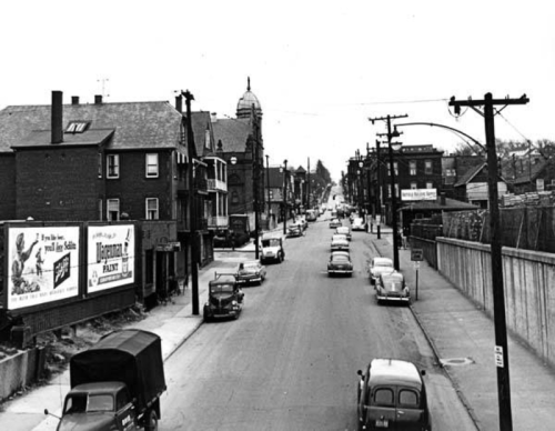Black and white photo of a busy street lined with buildings and vintage cars from the mid-20th century.