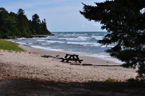 A serene beach scene with waves, a sandy shore, and a picnic table surrounded by trees under a clear sky.