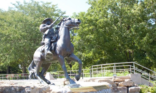 A bronze statue of a mounted soldier holding a rifle, set against a backdrop of trees and a stone base.
