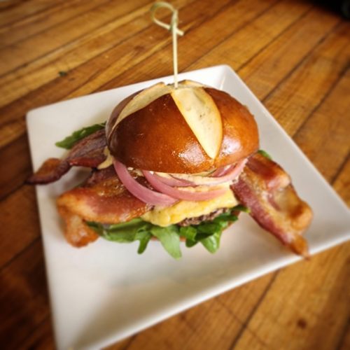 A pretzel bun sandwich with bacon, cheese, onions, and greens, served on a white plate.
