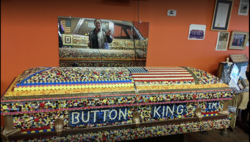 A colorful, decorative coffin adorned with buttons and an American flag, displayed in a room with orange walls.