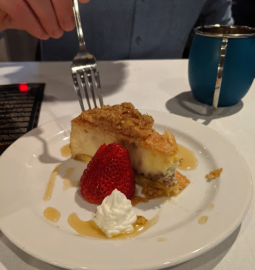 A slice of cheesecake topped with nuts, a strawberry, and whipped cream on a plate, with a fork in hand.