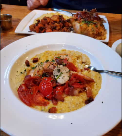 A bowl of creamy grits topped with shrimp, tomatoes, and bacon, served with a side of roasted vegetables.
