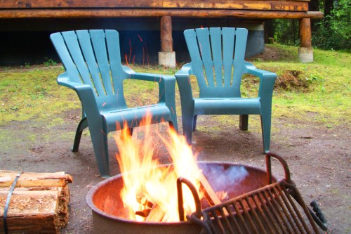 Two blue chairs beside a fire pit with flames, surrounded by logs, set in a natural outdoor setting.
