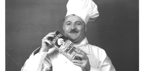 A smiling chef in a white uniform and hat holds a box of spaghetti dinner, posing for the camera.