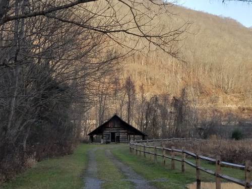 A rustic cabin along a gravel path, surrounded by bare trees and rolling hills in the background.