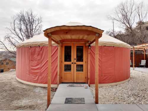 A round, tan yurt with a wooden door and a stone pathway, surrounded by bare trees and a cloudy sky.