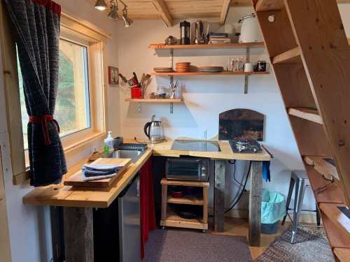 Cozy kitchen with wooden shelves, a sink, stove, and various kitchenware, under a staircase with a window view.