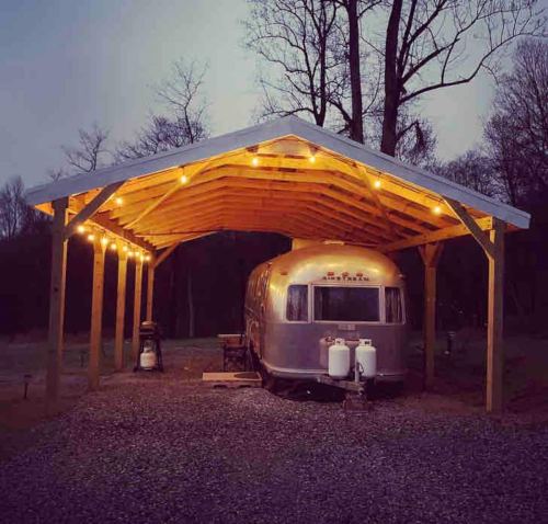 A silver Airstream trailer parked under a wooden shelter, illuminated by string lights at dusk.