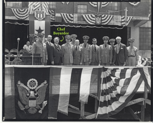 A group of men in military uniforms and suits stands on a stage decorated with flags and banners, labeled "Chef Boyardee."