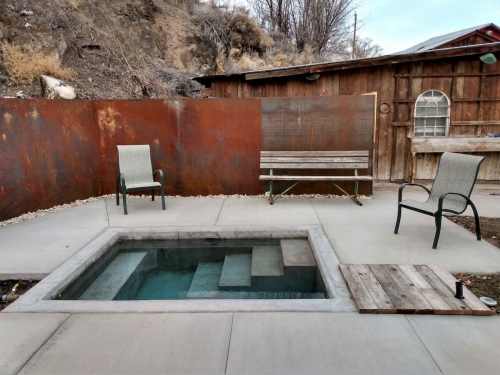 A small hot tub surrounded by concrete, with two chairs and a wooden bench against a rustic backdrop.