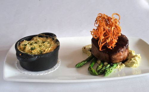 A gourmet dish featuring a filet mignon topped with crispy onions, asparagus, and a side of creamy gratin in a small dish.