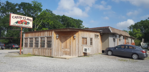 A rustic wooden building with a sign reading "Cajun Claws," surrounded by gravel and trees under a blue sky.
