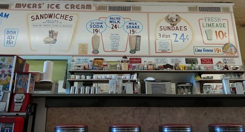 A vintage ice cream shop counter with menus for sandwiches, sodas, milkshakes, and sundaes displayed on the wall.