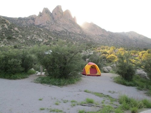 A colorful tent set up in a campsite surrounded by greenery, with mountains in the background and sunlight peeking over the peaks.