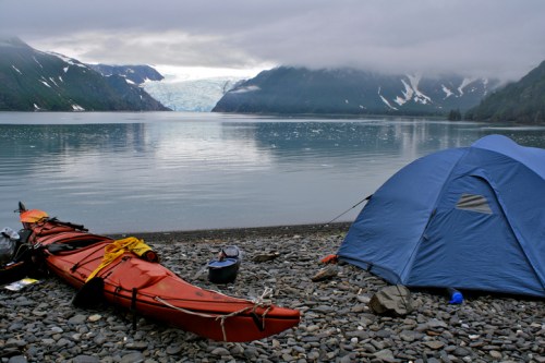 A red kayak rests on a rocky shore beside a blue tent, with a glacier and mountains in the background under cloudy skies.