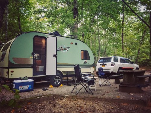 A green camper parked in a wooded campsite, with chairs and a cooler nearby, and a white SUV in the background.