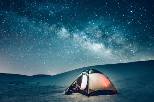 A tent illuminated at night under a starry sky filled with the Milky Way over a sandy landscape.