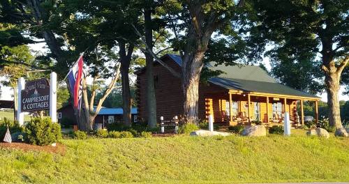 A rustic log cabin with a green roof, surrounded by trees and a flag, serving as a camp resort and cottages.