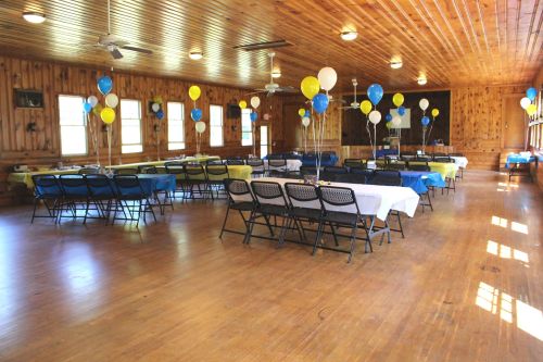 A spacious wooden hall set up for an event, with tables covered in blue and yellow linens and balloons.