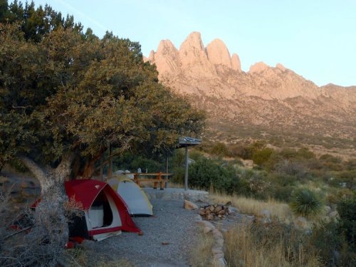 A campsite with red and yellow tents under a tree, set against a backdrop of rugged mountains at sunrise.