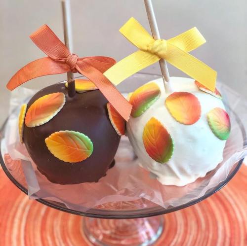 Two decorated caramel apples, one coated in dark chocolate with orange ribbon, the other in white chocolate with yellow ribbon, both adorned with colorful leaf-shaped candies.