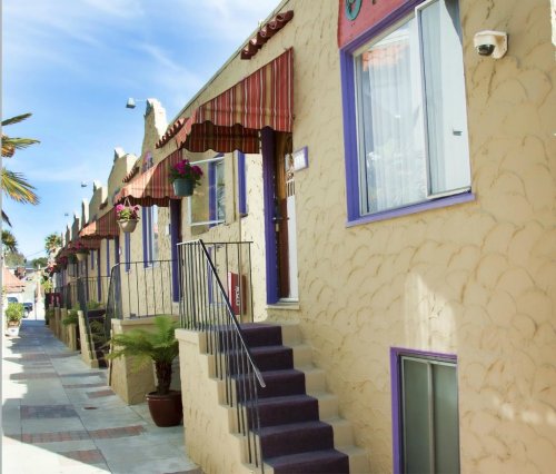 Colorful, textured building with stairs, awnings, and potted plants along a sunny sidewalk.