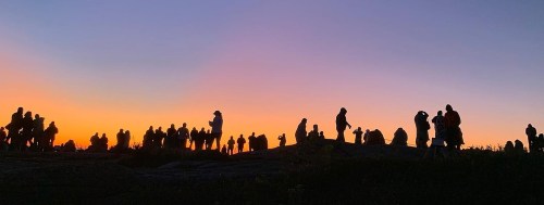 Silhouettes of people against a vibrant sunset, gathered on rocky terrain, creating a lively atmosphere.