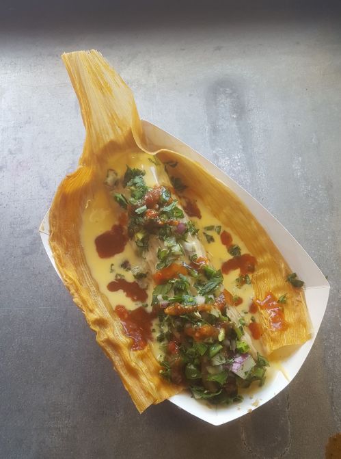 A tamale served in a corn husk, topped with cheese, salsa, and fresh herbs on a metal surface.