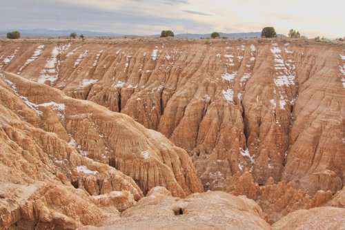 A rugged canyon landscape with steep, textured walls and patches of snow under a cloudy sky.