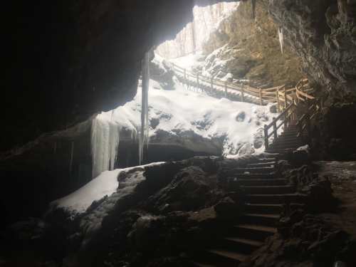 A snowy cave entrance with wooden stairs and icicles, leading to a bright, snowy landscape outside.
