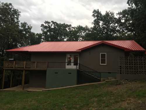 A modern house with a red metal roof, wooden deck, and surrounded by trees on a cloudy day.