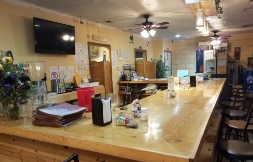 A cozy bar with a wooden counter, chairs, and a TV, featuring plants and decor in a warm, inviting atmosphere.