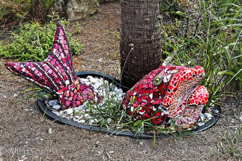 Colorful mosaic fish sculpture partially submerged in a garden bed, surrounded by pebbles and greenery.
