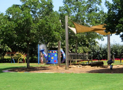A sunny park scene with a child playing near a colorful water feature and shaded areas under trees.