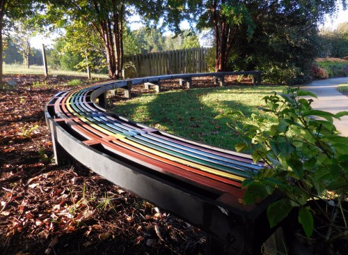 A curved, colorful bench surrounded by greenery in a park setting, with sunlight filtering through the trees.