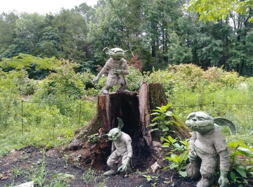 Three whimsical goblin sculptures are positioned around a tree stump in a lush, green garden setting.
