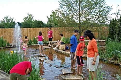 Children and adults enjoy a playful day by a pond with stepping stones, surrounded by greenery and a wooden fence.