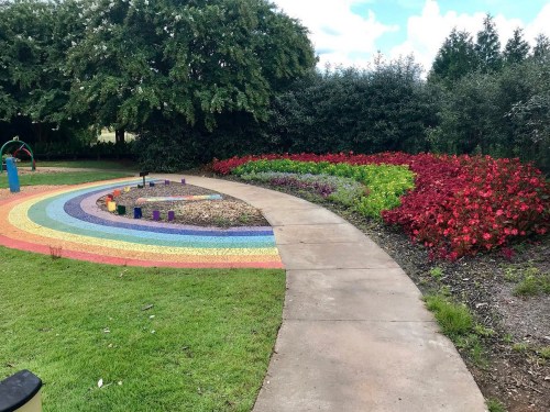 A colorful spiral path made of stones leads through a garden with vibrant flowers and greenery under a blue sky.