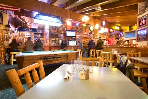A cozy bar scene with wooden decor, a pool table, and patrons enjoying drinks at the counter. Warm lighting creates a welcoming atmosphere.