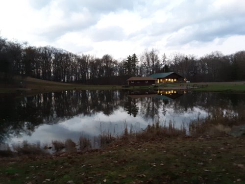 A serene pond reflects a cabin surrounded by trees under a cloudy sky at dusk.