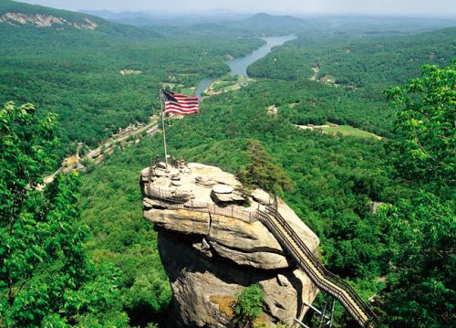 Aerial view of a rocky outcrop with an American flag, surrounded by lush green mountains and a winding river below.