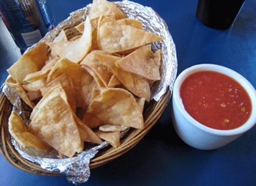 A basket of crispy tortilla chips next to a small bowl of red salsa on a blue table.