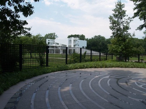 A modern white building is visible behind a circular stone pathway, surrounded by greenery and trees under a cloudy sky.